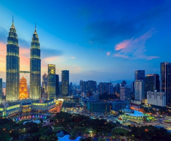 Panoramic view of a modern city skyline at dusk, featuring the illuminated Petronas Twin Towers, surrounded by numerous skyscrapers and a park with lush trees and water features, under a vibrant blue and pink sky.