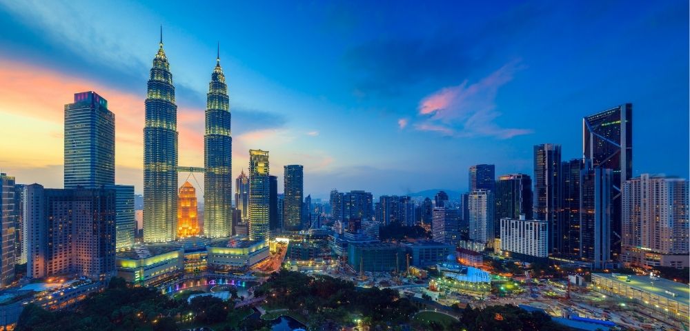 Panoramic view of a modern city skyline at dusk, featuring the illuminated Petronas Twin Towers, surrounded by numerous skyscrapers and a park with lush trees and water features, under a vibrant blue and pink sky.
