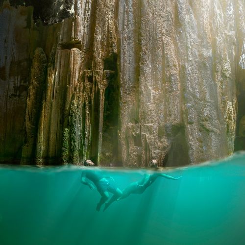 Split-level view of a swimmer gliding underwater in turquoise water beneath towering, sunlit rock walls, creating a serene, adventurous mood.