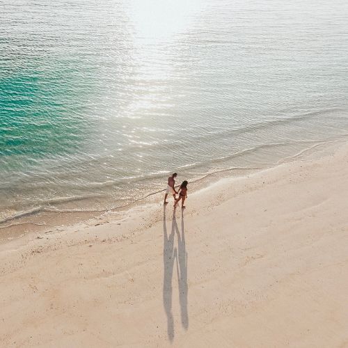Aerial view of two people walking hand in hand along a sunlit sandy beach, with long shadows, gentle waves, and calm turquoise water.