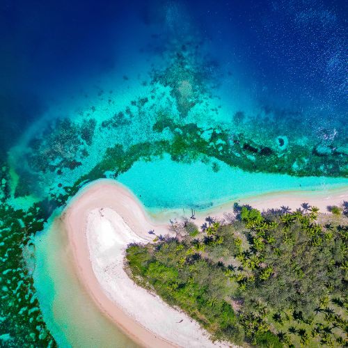 Aerial view of a tropical beach and turquoise lagoon, with white sand curving around a lush, palm-lined island and shallow coral reefs.