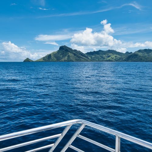 View from a boat across deep blue ocean toward a lush, mountainous tropical island beneath bright clouds; metal bow railing frames the serene scene.
