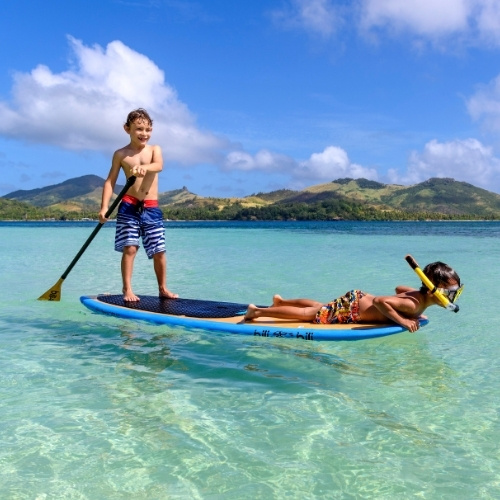 Smiling boys paddleboard on a tropical lagoon; clear turquoise water, green mountains, and blue skies create a playful scene.