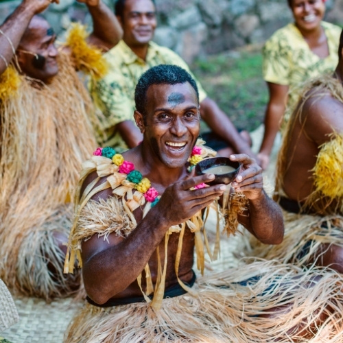Smiling man in a traditional straw outfit holds a bowl during a lively cultural celebration, surrounded by cheering people in a lush setting.