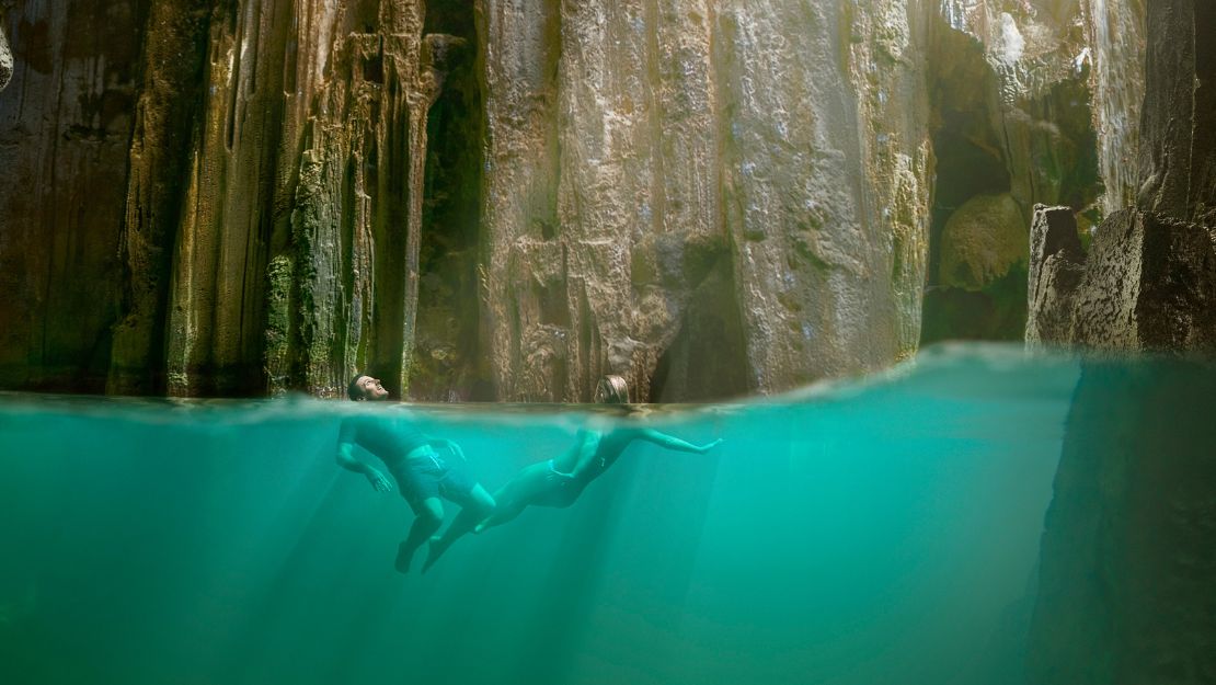 Split-level view of a swimmer gliding underwater in turquoise water beneath towering, sunlit rock walls, creating a serene, adventurous mood.