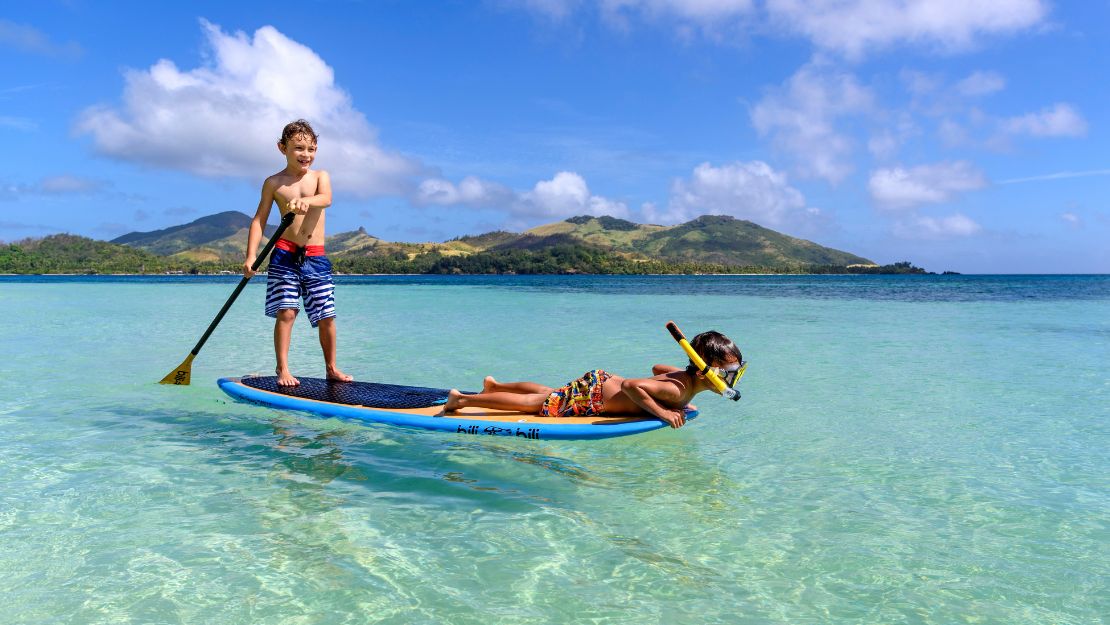 Smiling boys paddleboard on a tropical lagoon; clear turquoise water, green mountains, and blue skies create a playful scene.