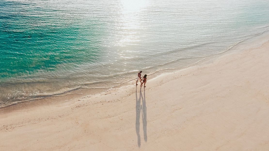 Aerial view of two people walking hand in hand along a sunlit sandy beach, with long shadows, gentle waves, and calm turquoise water.