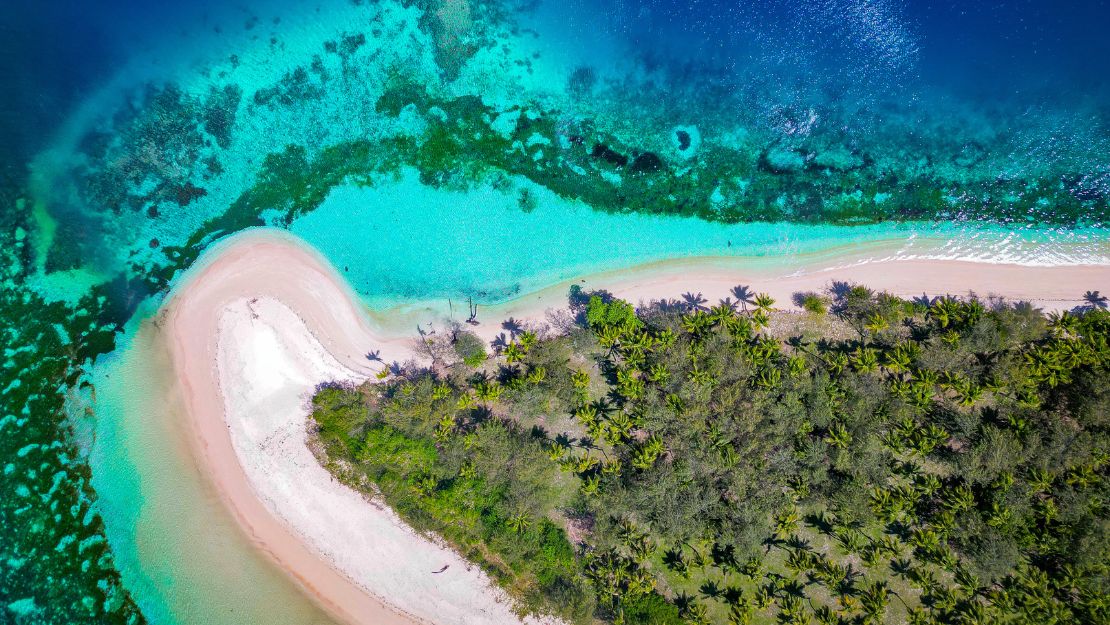Aerial view of a tropical beach and turquoise lagoon, with white sand curving around a lush, palm-lined island and shallow coral reefs.