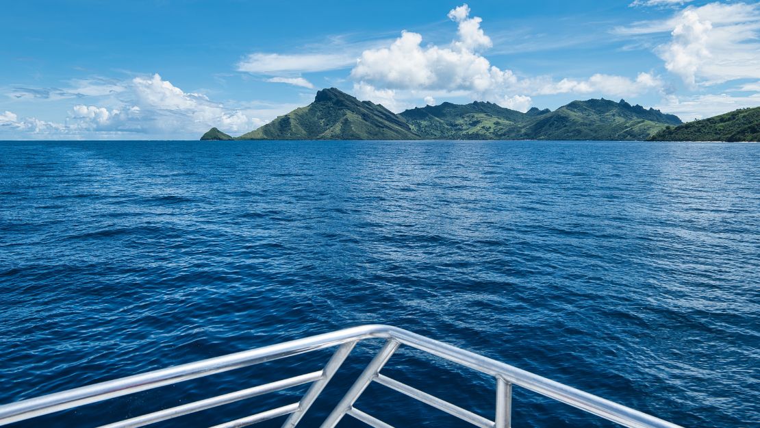 View from a boat across deep blue ocean toward a lush, mountainous tropical island beneath bright clouds; metal bow railing frames the serene scene.