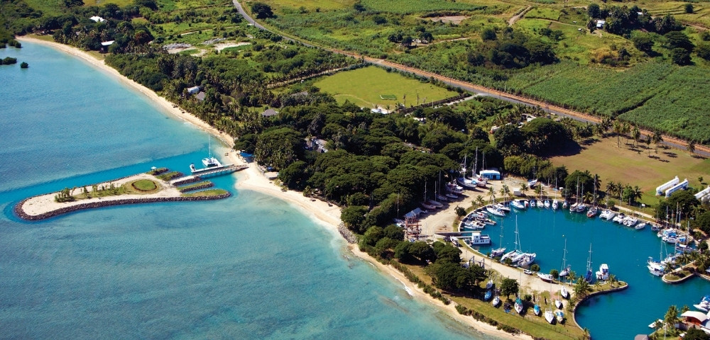 Aerial view of a tropical coastal marina with sailboats, turquoise water, sandy beaches, lush fields, and a peaceful resort setting.