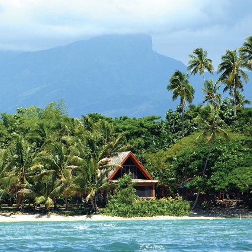 Tropical island beach with turquoise water, swaying palms, and a rustic cabin beneath a dramatic misty mountain; serene escape.