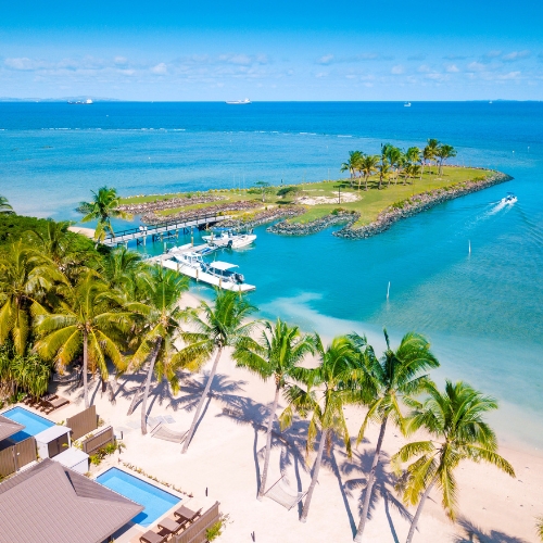 Aerial view of a tropical beach resort with swaying palms, white sand, turquoise lagoon, marina, boats, and a tranquil breakwater.