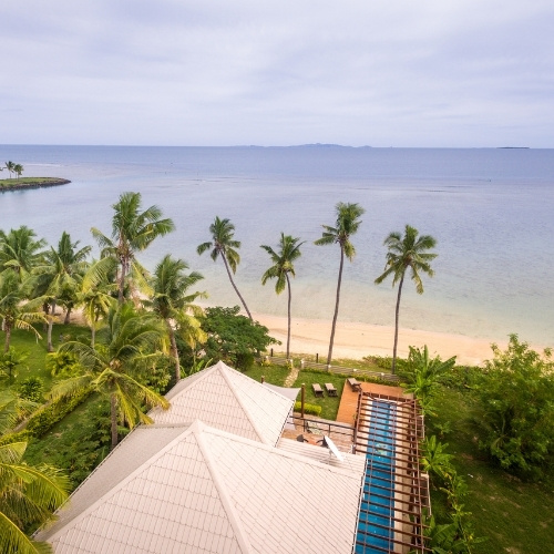 Tropical beachfront resort with white-roofed buildings, swaying palm trees, sandy beach, and calm blue ocean beneath an overcast sky.