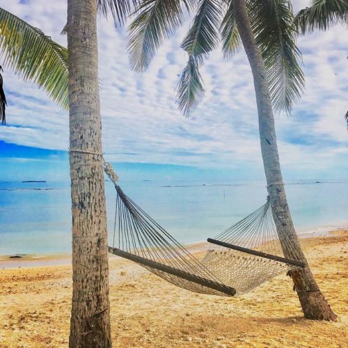 A woven hammock hangs between two palm trees on a sandy tropical beach, overlooking calm turquoise ocean beneath a bright, cloudy sky.