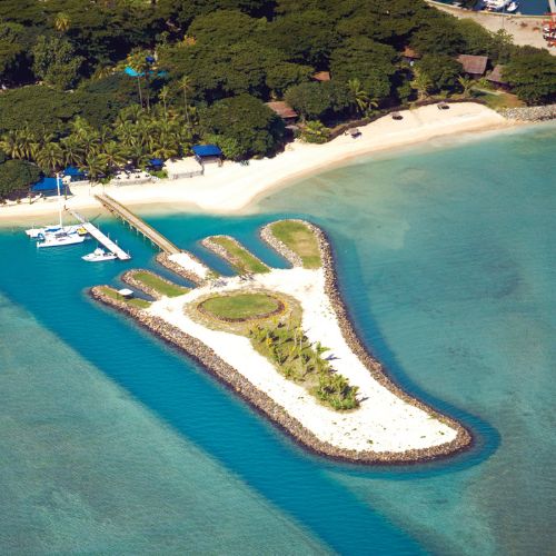 Aerial view of a tropical island resort with white sandy beaches, turquoise lagoons, lush palms, a marina, and a peaceful landscaped islet.