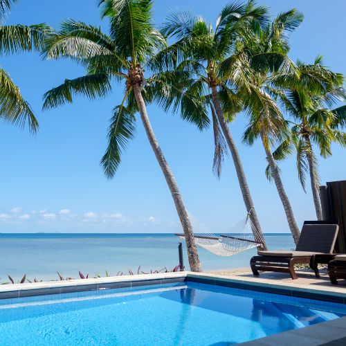 Poolside view with palm trees, a hammock, and lounge chairs overlooking a calm ocean under a clear blue sky.