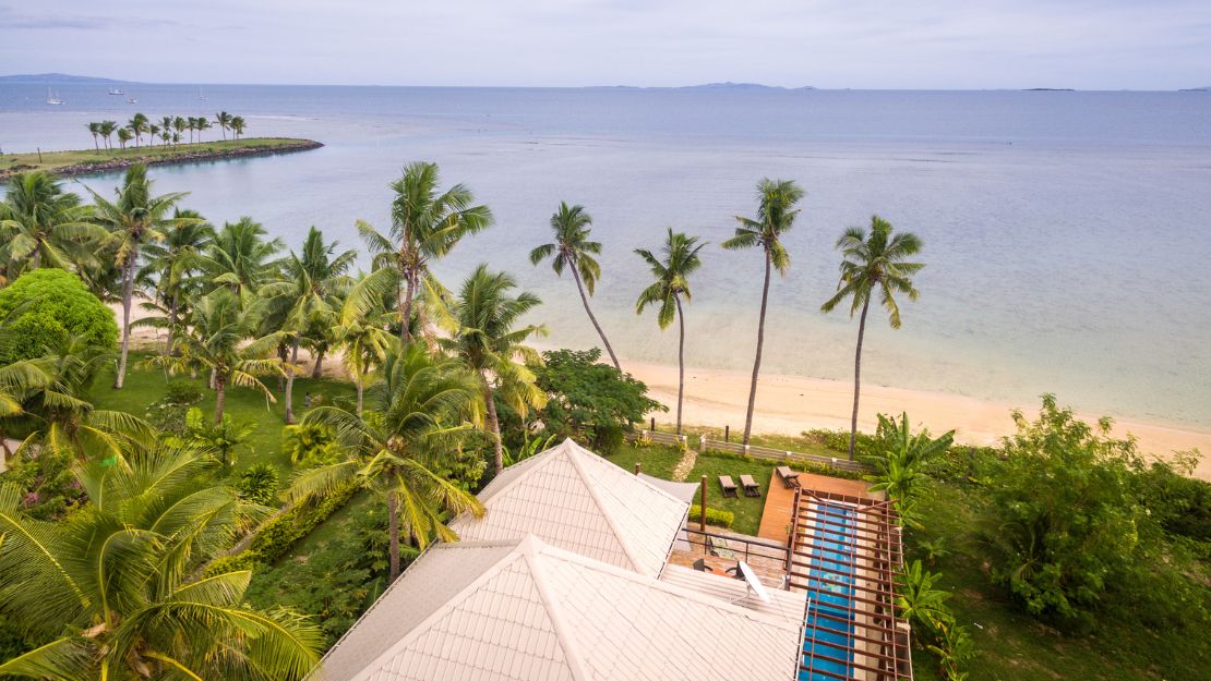 Tropical beachfront resort with white-roofed buildings, swaying palm trees, sandy beach, and calm blue ocean beneath an overcast sky.