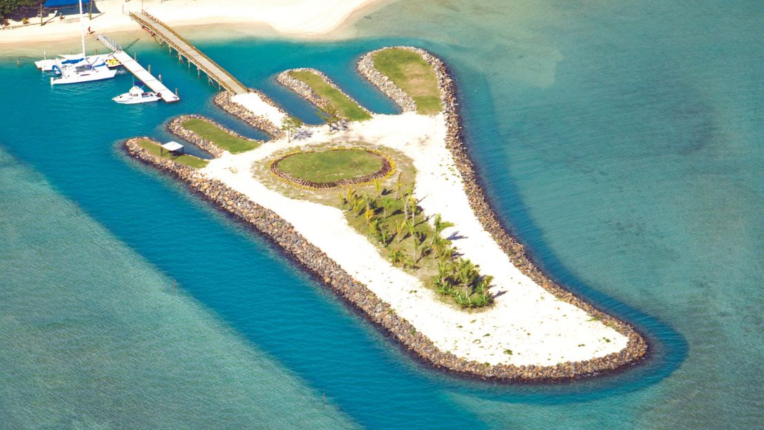 Aerial view of a tropical island resort with white sandy beaches, turquoise lagoons, lush palms, a marina, and a peaceful landscaped islet.