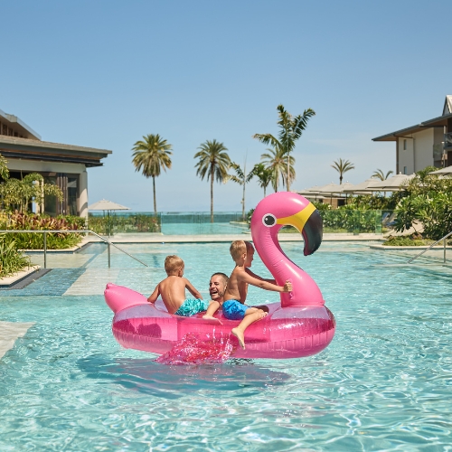 Children laugh and play on a pink flamingo float in a resort pool, with palm trees, ocean views, and bright sunshine creating a joyful mood.