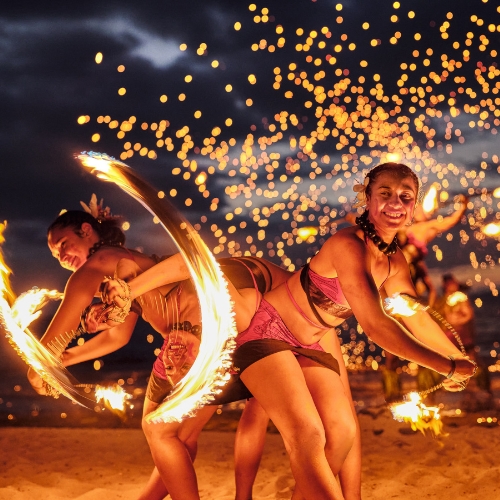 Two smiling fire dancers spin flaming hoops on a beach at night, scattering glowing sparks and creating an energetic, festive scene.