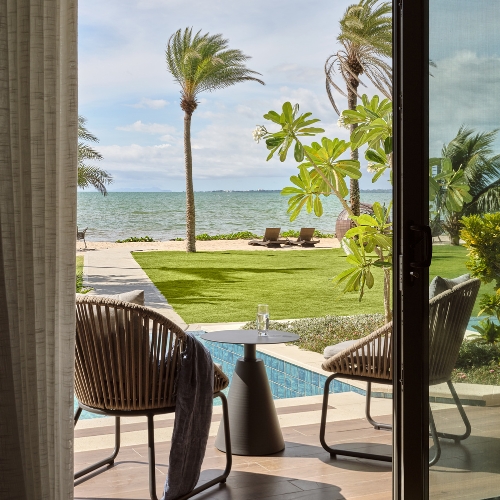 Open patio doors frame a serene tropical beachfront view with palm trees, turquoise sea, pool, green lawn, lounge chairs, and seating.