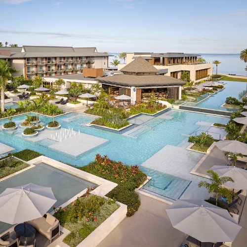 Aerial view of a luxurious seaside resort with interconnected turquoise pools, shaded cabanas, tropical gardens, modern buildings, and ocean beyond.