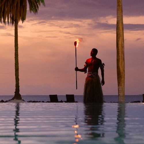 Silhouetted person in a grass skirt holds a flaming torch behind a tropical infinity pool at sunset, framed by palm trees and calm ocean.