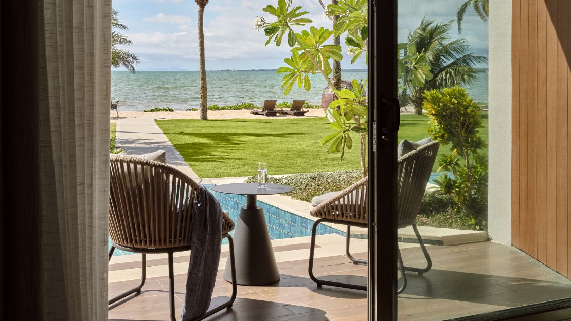 Open patio doors frame a serene tropical beachfront view with palm trees, turquoise sea, pool, green lawn, lounge chairs, and seating.