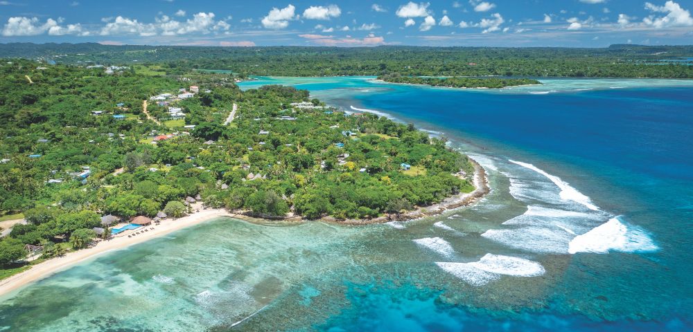 Aerial view of a lush, green tropical island with a white sand beach, clear turquoise water revealing coral reefs, and deep blue ocean under a cloudy sky.