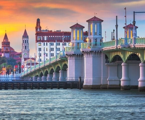 Historic bridge with ornate towers spanning a body of water at sunset, with a city skyline in the background.