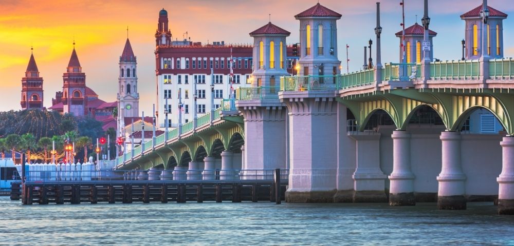 Historic bridge with ornate towers spanning a body of water at sunset, with a city skyline in the background.