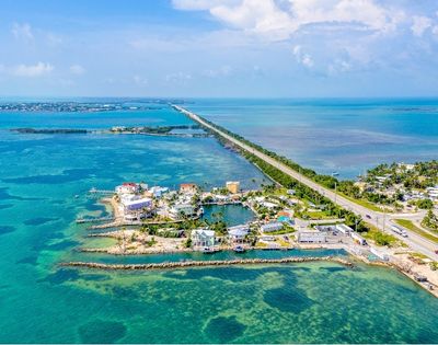 Aerial view of a causeway stretching across turquoise waters, connecting a coastal town with brightly colored buildings and marinas to a distant horizon.