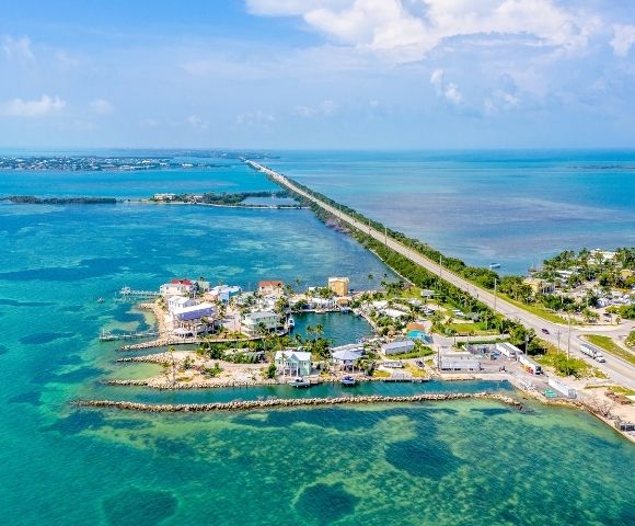 Aerial view of a causeway stretching across turquoise waters, connecting a coastal town with brightly colored buildings and marinas to a distant horizon.
