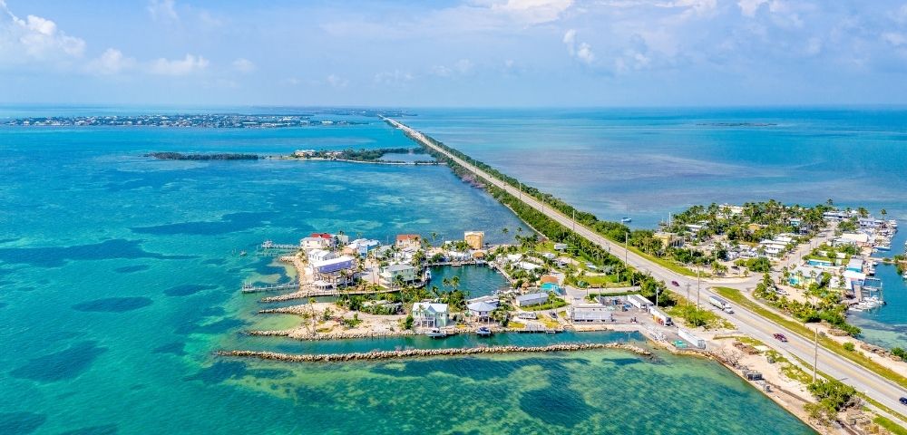 Aerial view of a causeway stretching across turquoise waters, connecting a coastal town with brightly colored buildings and marinas to a distant horizon.