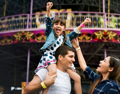 A young girl rides on her father's shoulders with her arms raised in excitement at an amusement park, while her mother reaches up towards her.