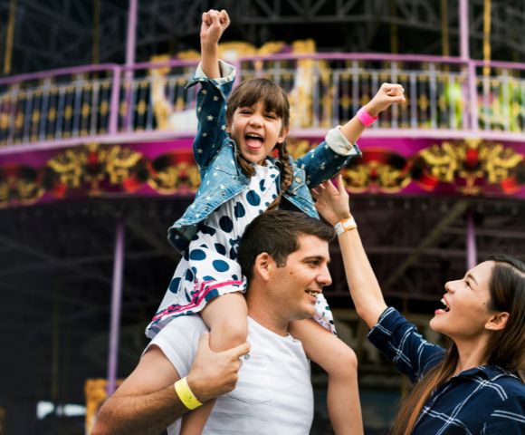 A young girl rides on her father's shoulders with her arms raised in excitement at an amusement park, while her mother reaches up towards her.