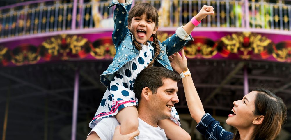 A young girl rides on her father's shoulders with her arms raised in excitement at an amusement park, while her mother reaches up towards her.