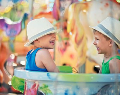 Two young boys wearing straw hats laugh heartily while sitting in a carousel enclosure, with a colorful, blurred carousel background.