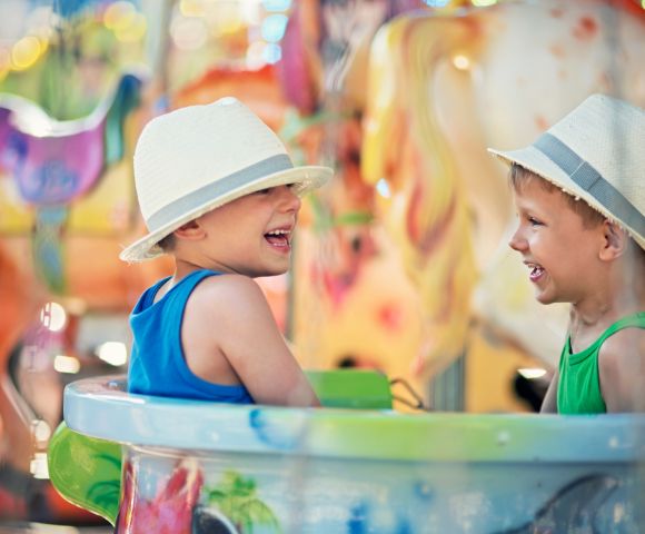 Two young boys wearing straw hats laugh heartily while sitting in a carousel enclosure, with a colorful, blurred carousel background.