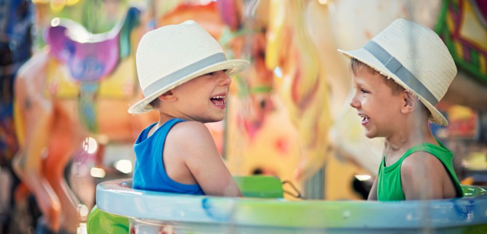 Two young boys wearing straw hats laugh heartily while sitting in a carousel enclosure, with a colorful, blurred carousel background.