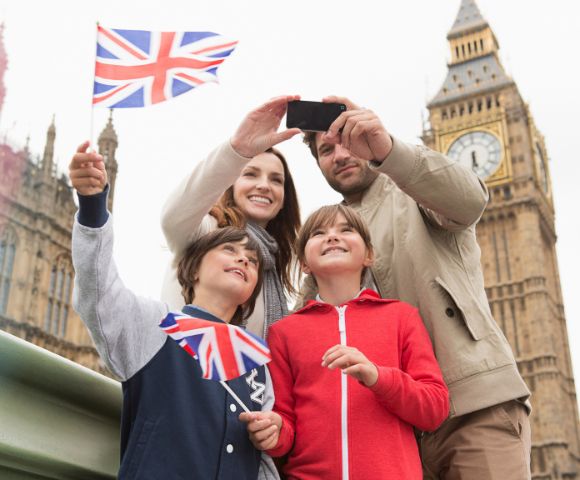 A family takes a selfie with Big Ben and the Houses of Parliament in the background under a Union Jack flag.