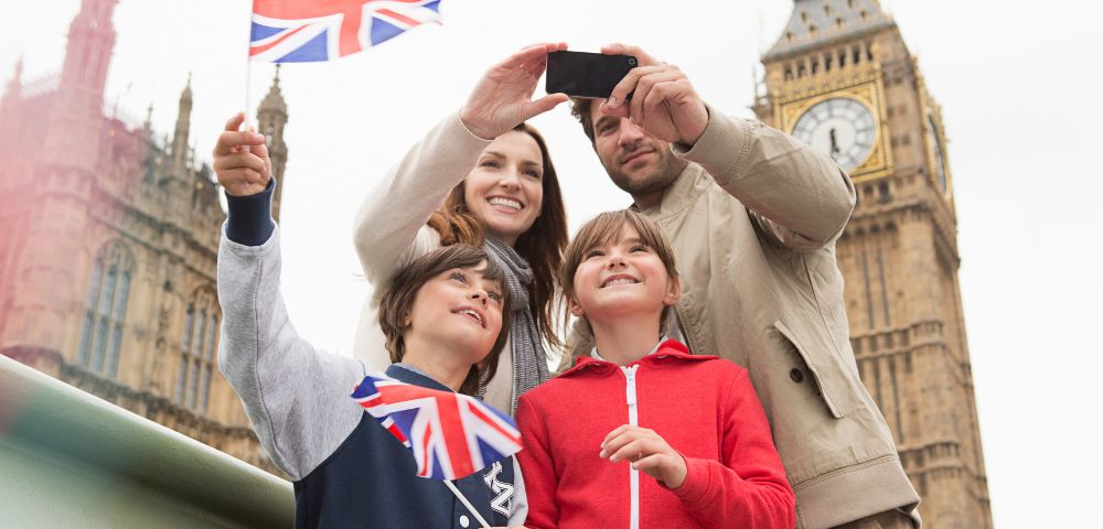 A family takes a selfie with Big Ben and the Houses of Parliament in the background under a Union Jack flag.