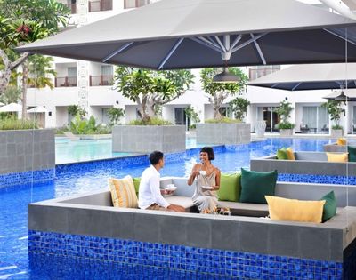 A couple enjoys drinks while seated in a sunken lounge area next to a bright blue swimming pool, under a large gray umbrella at a resort.