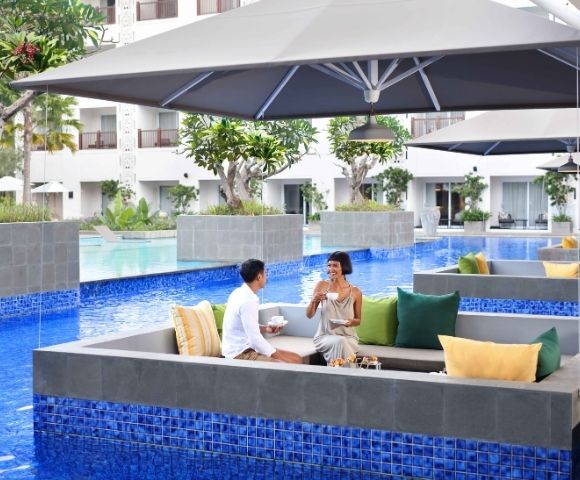 A couple enjoys drinks while seated in a sunken lounge area next to a bright blue swimming pool, under a large gray umbrella at a resort.