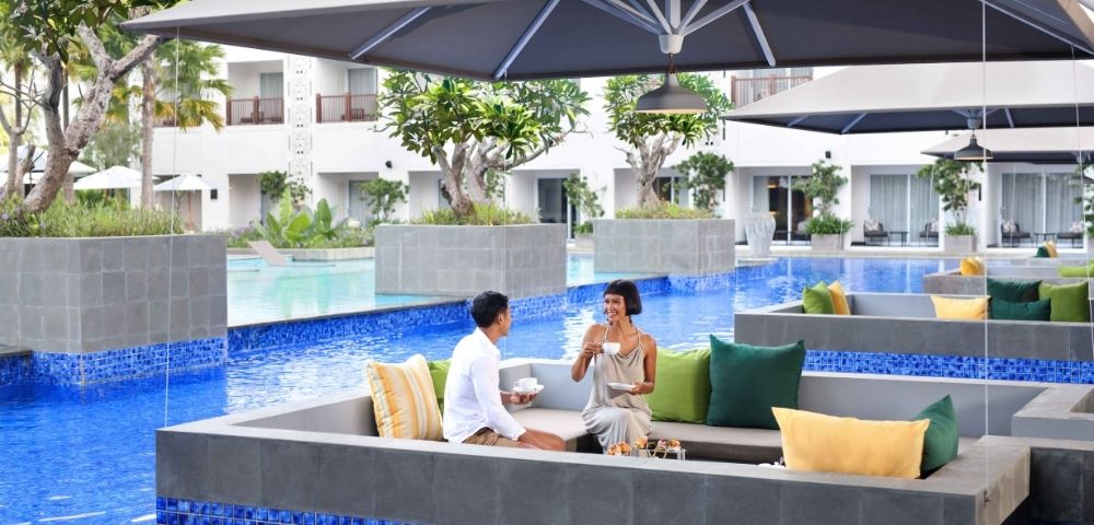 A couple enjoys drinks while seated in a sunken lounge area next to a bright blue swimming pool, under a large gray umbrella at a resort.
