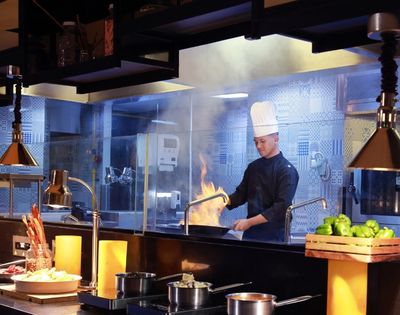 Chef in a tall white hat cooks with flames erupting from a pan in a professional kitchen.
