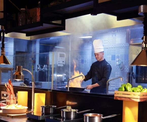 Chef in a tall white hat cooks with flames erupting from a pan in a professional kitchen.