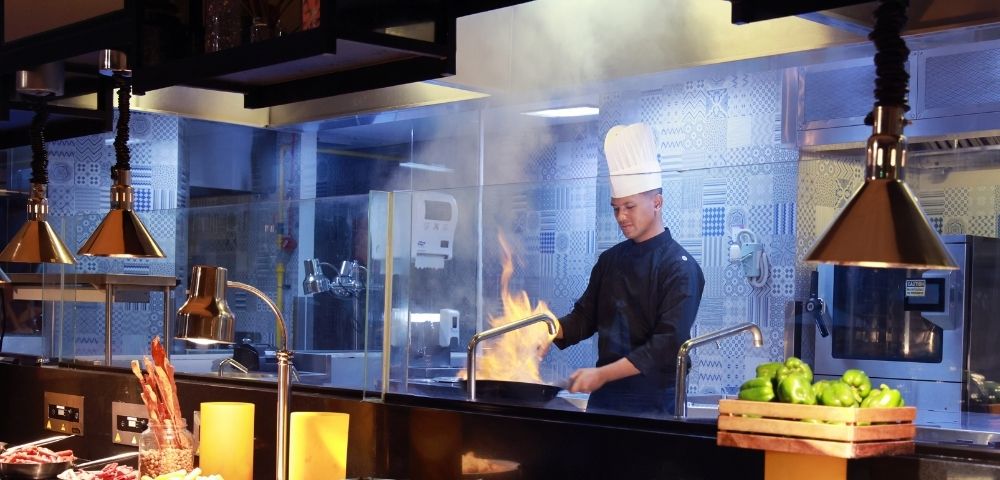 Chef in a tall white hat cooks with flames erupting from a pan in a professional kitchen.