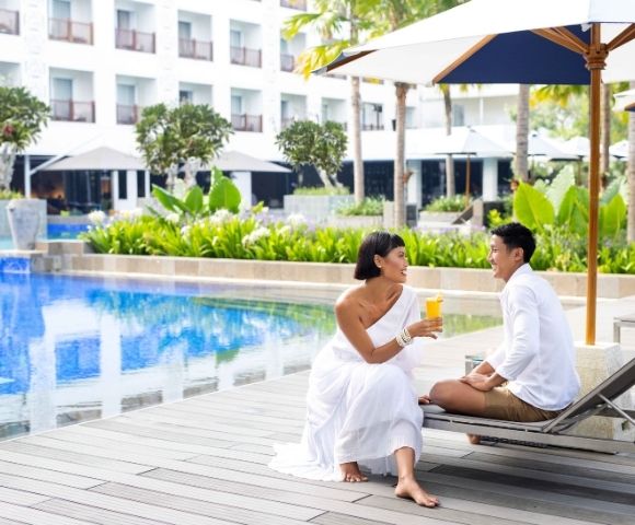 A couple sits poolside, with one person holding a drink and facing the other, at a resort with a white hotel building in the background.
