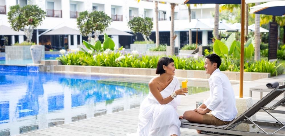 A couple sits poolside, with one person holding a drink and facing the other, at a resort with a white hotel building in the background.